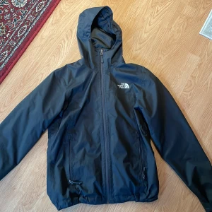 Svart vindjacka från The North Face - Säljer en svart vindjacka från The North Face i mycket bra skick. Jackan har en dragkedja framtill och en justerbar huva. Perfekt för höst och vår, och den är långärmad med en normal passform. Den är både stilren och funktionell för vardagsbruk.