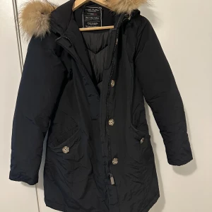 Svart parkajacka med pälsdetaljer - Säljer en snygg svart parkajacka med äktapäls på huvan. Jackan har långa ärmar och stängs med både dragkedja och knappar. Perfekt för kalla vinterdagar och höstpromenader. Den har praktiska fickor framtill och en skön passform. Ett måste för garderoben!