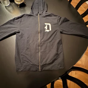 Svart hoodie från Dondup - Säljer en svart hoodie från Dondup Kids i mycket bra skick. Den har en dragkedja framtill och en stor vit bokstav 'D' på bröstet. Den e i största storleken i kids och passar nästan mig som e 168cm den e bara lite kort för mig i armarna, så att den är stor för att vara i kids, skriv vid intresse och frågor, pris är ej hugget i sten🤩