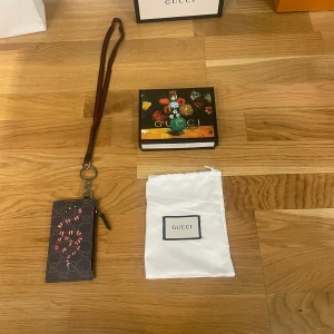 Gucci plånbok med nyckelring - Säljer en snygg Gucci plånbok i svart skinn med mönster av ormar. Den har en praktisk dragkedja och en nyckelring med rem för enkel användning. Perfekt för att hålla dina kort och småsaker säkra. Kommer med originalförpackning och påse.