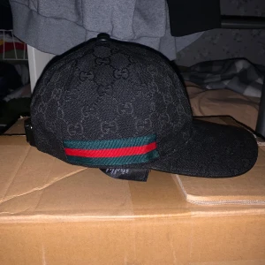 Gucci keps  - Säljer en snygg svart keps från Gucci med det klassiska mönstret och en grön-röd randdetalj. Kepsen är i storlek M och har justerbar rem baktill. Perfekt för att ge din outfit en lyxig touch!