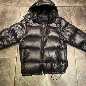 Svart pufferjacka från Polo Ralph Lauren - Säljer en snygg svart pufferjacka från Polo Ralph Lauren i storlek XL. Jackan är riktigt varm, perfekt för kalla dagar. Den är mycket sparsamt använd och ser i princip ny ut. Inga märken eller liknande. Perfekt för vintern! Fler bilder kan ordnas och några hundralappar billigare vid snabb affär.