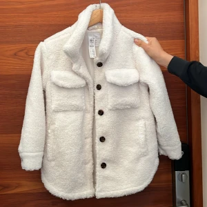 Vit teddyjacka - Supermysig vit teddyjacka med stora knappar och två bröstfickor. Perfekt för kyliga dagar när du vill vara både varm och stilren. Jackan har en skön och fluffig känsla, vilket gör den idealisk för höst och vinter. Passar perfekt för en casual look eller när du bara vill mysa till det lite extra.