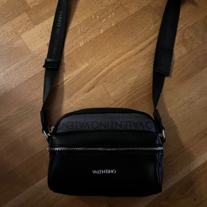 Svart axelväska från Valentino - Snygg svart axelväska från Valentino i mycket bra skick. Väskan har en justerbar axelrem med logotyp och en praktisk dragkedja upptill. Perfekt för både vardag och fest! 🖤