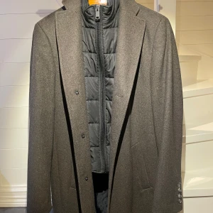 Wool-coat from Duffel & Rums - Original price 300 euro  	•	Lapel collar 	•	Removable vest insert with stand-up collar 	•	Full-length button placket  •	2 slanted side pockets 	•	2 inner pockets 	•	Cuff buttons 	•	Back vent  The wool coat from Duffel & Rums impresses with its elegant and versatile design. The vest insert can be quickly removed as desired, making the coat adaptable enough to suit everything from formal to sporty occasions. Just the right length to cover a blazer.