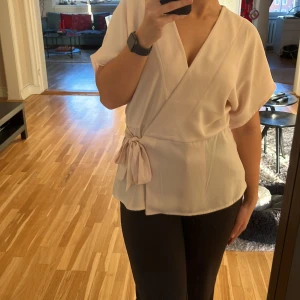 Ljusrosa omlottopp från Vila - Säljer en superfin ljusrosa omlottblus från Vila i storlek 38 då den är lite stor för mig. Toppen har korta ärmar och ett knytband i midjan som ger en snygg passform. Perfekt för både vardag och fest! ✨