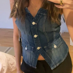 Jeansväst med knappar - Snygg jeansväst i mörkblå denim med knappar framtill. Västen har en v-ringad design och två fickor som ger en cool look. Passar både till vardags och fest!