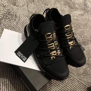 Svarta sneakers från Versace Jeans Couture - Snygga svarta sneakers från Versace Jeans Couture med guldtext på ovansidan. De har en modern design med snörning och är perfekta för både vardag och fest. Skorna är i nytt skick och kommer i originalförpackning.