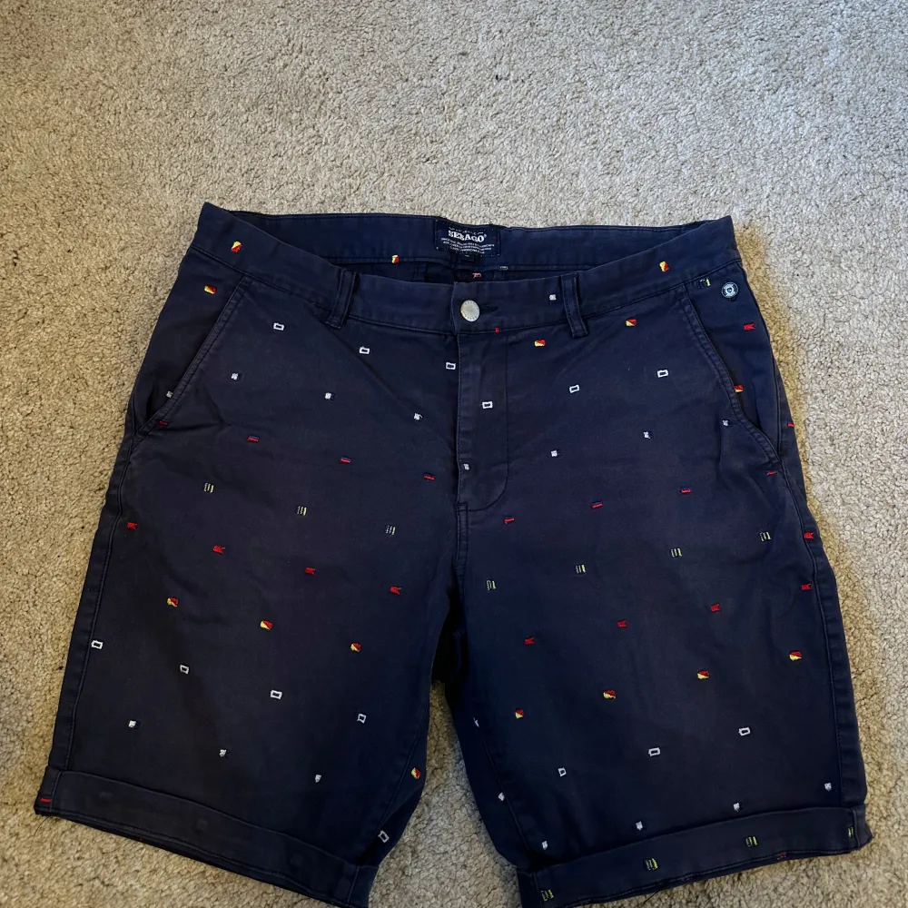 Säljer ett par mörkblå shorts från Sebago i mycket bra skick. De har ett coolt broderat mönster i olika färger som ger en unik look. Perfekta för sommaren med en bekväm passform och stilren design. Passar både till vardags och festliga tillfällen! Den är solskadad men inget som syns i bruk.. Shorts.