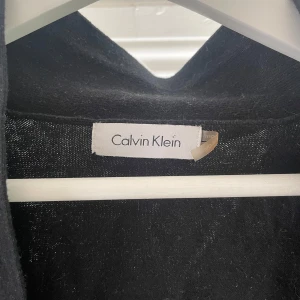 Svart bolero från Calvin Klein - Snygg svart bolero från Calvin Klein i ett elegant och stilrent snitt. Perfekt för att lägga till en touch av klass till vilken outfit som helst. Den är långärmad och passar bra till både vardag och fest. Ett måste i garderoben för den modeintresserade!