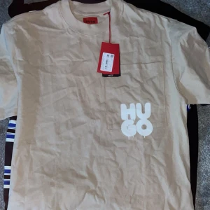 Beige t-shirt från Hugo - Säljer en snygg beige t-shirt från Hugo i nytt skick. Den har en cool vit logga på bröstet och en liten ficka. Perfekt för en avslappnad stil och passar till både vardag och fest. Storleken är normal och materialet känns mjukt och skönt.