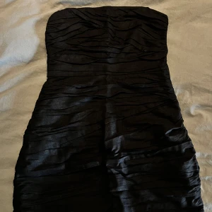 Svart axelbandslös klänning från Zara - Snygg svart axelbandslös klänning från Zara i ett elegant och stilrent utförande. Klänningen har en tight passform och är perfekt för festliga tillfällen. Den är tillverkad i ett glansigt material och har en dragkedja i sida för enkel påklädning. 