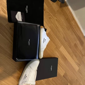 Vita sneakers från Saint Laurent - Säljer ett par stilrena vita sneakers från Saint Laurent. De har en klassisk design med svart detalj vid hälen och Saint Laurent-logga på sidan. Skorna är i bra skick med lite slitage på sulorna. Perfekta för både vardag och fest! Skriv gärna om du har frågor och funderingar tveka inte och skriv pris kan diskuteras!!!