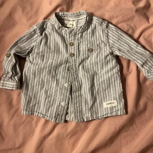 Newbie skjorta  - Säljer en superfin randig skjorta från Newbie i storlek 62. Skjortan är i ett mjukt bomullsmaterial med knappar framtill och en liten bröstficka. Den har långa ärmar och en klassisk krage. Använd ksk 2 ggr.