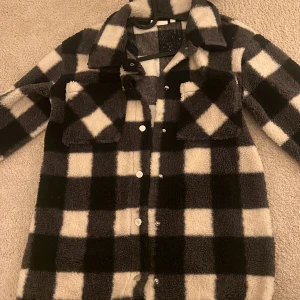 Svartvit rutig teddyjacka - Supermysig svartvit rutig teddyjacka i oversized stil. Perfekt för höst och vinter med sina långa ärmar och knappar framtill. Jackan har två stora fickor på bröstet och är gjord i ett mjukt och varmt material. Perfekt för att hålla sig varm och stilren under kyliga dagar!