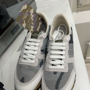 Valentino Garavani sneakers i grått och vitt - Säljer ett par snygga Valentino Garavani sneakers i grått och vitt med coolt camouflage-mönster. Skorna har snörning och är i nytt skick. Perfekta för både vardag och fest! Kommer i originalförpackning.