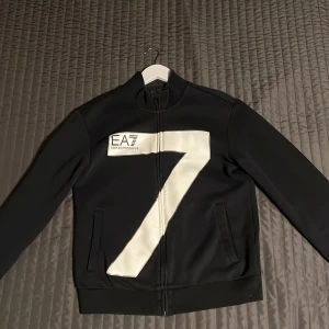 Svart jacka från Emporio Armani - Säljer en svart hoodie   från Emporio Armani med en stor vit siffra 7 på framsidan. Hoodien har dragkedja och är långärmad, perfekt för en stilren look. Den är i mycket bra skick och passar  till vardags.