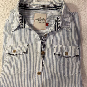 Randig skjorta från Redgreen - Säljer en snygg blå- och vitrandig skjorta från Redgreen. Den har långa ärmar och två bröstfickor med knappar. Skjortan är i bomull och har en klassisk krage. Perfekt för både vardag och lite mer uppklädda tillfällen. Passar bra till jeans för en avslappnad look.