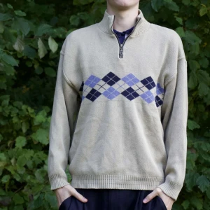 Beige tröja med argylemönster - Säljer en snygg beige tröja med klassiskt argylemönster i blått och lila. Tröjan har en dragkedja vid halsen och är perfekt för höst och vinter. Den är långärmad och har ribbade muddar vid ärmslut och nederkant. Perfekt för en stilren och bekväm look!