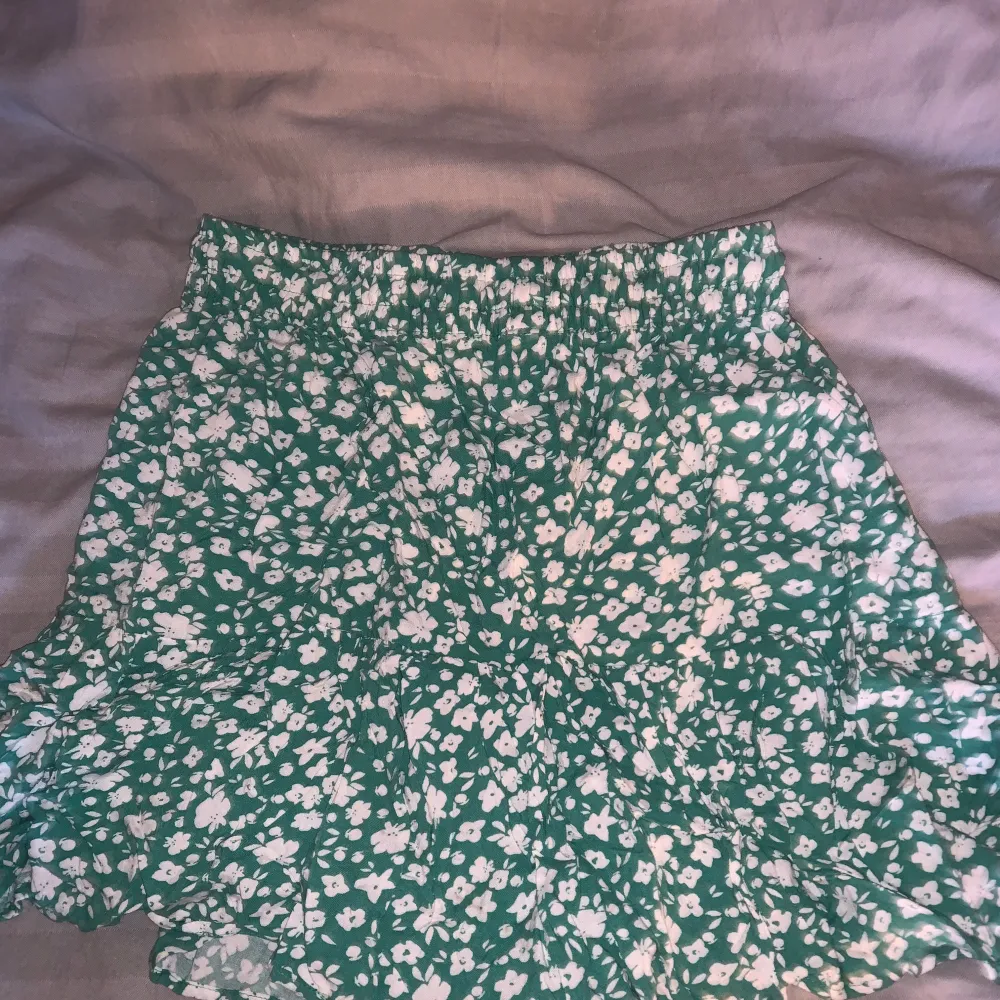 Säljer en superfin grön kjol med vita blommor. Det finns även inbyggda shorts!! Den är perfekt för sommaren med sitt lätta och luftiga material. Kjolen har en elastisk midja för en bekväm passform och en söt volangdetalj längst ner. Perfekt för en dag på stranden eller en somrig picknick! 🌸. Kjolar.