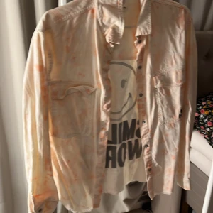 Vit och orange skjorta med Smiley World-tryck - Cool vit skjorta med orange batikmönster och stort Smiley World-tryck på ryggen. Skjortan har långa ärmar och knappar framtill. Perfekt för en avslappnad stil med en touch av färg och humor.