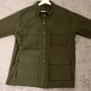 Grön tjockare skjorta Gina Tricot - Säljer en grön skjortjacka från Gina Tricot med knappar framtill och fyra stora fickor. Perfekt för lager-på-lager-stil. Den har en klassisk krage och är tillverkad i ett slitstarkt material.