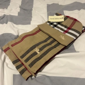 Beige rutig mössa från Burberry - Snygg beige mössa från Burberry med klassiskt rutigt mönster i svart, rött och vitt. Mössan är broderad med Burberrys logotyp och är perfekt för att hålla dig varm med stil.