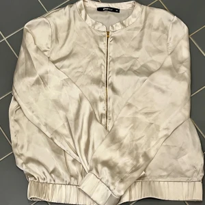 Beige bomberkofta från Gina Tricot - Snygg beige bomberkofta från Gina Tricot i ett glansigt material. Koftan har en dragkedja i guld och långa ärmar. Fläck på framsidan. 