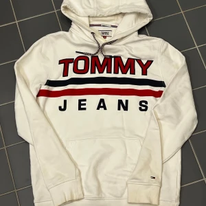 Vit hoodie från Tommy Jeans - Säljer en snygg vit hoodie från Tommy Jeans med stora röda och blå bokstäver på framsidan. Hoodien har en justerbar huva med snören i rött, vitt och blått. Perfekt för en casual look!