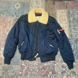 Blå bomberjacka från Alpha Industries - Säljer en snygg blå bomberjacka från Alpha Industries med en mjuk, beige pälsliknande krage. Jackan har en dragkedja framtill och en cool detalj med 'Remove Before Flight'-tag på ärmen. Perfekt för höst och vinter!