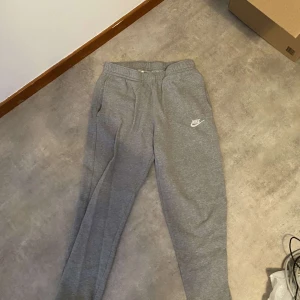 Grå mjukisbyxor från Nike - Säljer ett par grå mjukisbyxor från Nike med elastisk midja och sidofickor. Perfekta för en avslappnad stil. Nike-loggan är tryckt på ena benet.