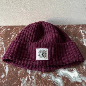 Stone Island mössa - Snygg vinröd mössa från Stone Island i ribbat material. Mössan har en uppvikt kant med en broderad logotyp framtill. Perfekt för att hålla värmen med stil. Skicket är bra och inga hål eller defekter fins, endast små tecken på användning. Priset är inte hugget i sten. Tveka inte att skriva vid frågor😄.
