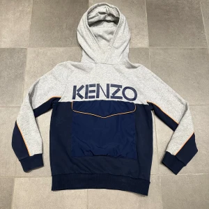 Grå och marinblå hoodie från Kenzo - Storlek: 14 år | Säljer en snygg grå och marinblå hoodie från Kenzo med stor logga på bröstet. Tröjan har en stor ficka framtill med orange detaljer och en bekväm huva. Perfekt för en stilren och avslappnad look.