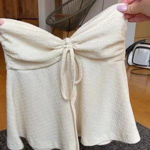 Beige axelbandslös topp från Shein - Säljer en söt beige axelbandslös topp från Shein. Toppen har en knytning framtill och är tillverkad i ett mjukt, texturerat material. Perfekt för varma dagar eller som en del av en lager-på-lager look.