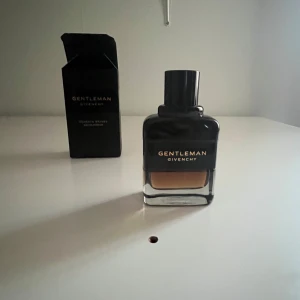 Gentleman Givenchy - Elegant herrparfym från Givenchy i en stilren flaska med svart lock och etikett. Doften är klassisk och sofistikerad, perfekt för den moderna gentlemannen. Flaskan har en fyrkantig form med en transparent del som visar parfymens gyllene färg. Saknar någon enstaka ml. 60 ml.