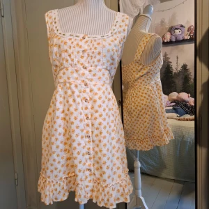 Blommig sommarklänning med volang - Charmig vit klänning med små gula blommor och volangdetaljer. Klänningen har en söt spetskant vid halsringningen och knappar framtill. Perfekt för sommarens varma dagar!