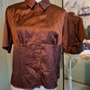 Brun kortärmad blus - Snygg brun blus med korta ärmar och knappar framtill. Perfekt för en stilren look. Tillverkad i ett glansigt material som ger en elegant touch. Lappen finns tyvärr inte kvar så vet inte vad märket eller storleken är. Uppskattas till en L
