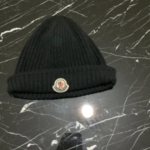 Svart ribbad mössa från Moncler - Snygg svart ribbad mössa från Moncler med broderad logotyp framtill. Perfekt för kyliga dagar och ger en stilren look. Tillverkad i mjukt material för extra komfort. ( man kan pruta )