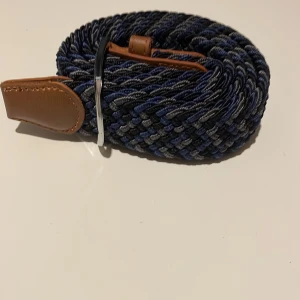 Flätat bälte  - Snyggt flätat bälte i blå och grå toner med bruna skinn-detaljer. Perfekt för att ge en stilren touch till din outfit. Bältet har en silverfärgad spänne som ger en modern känsla.