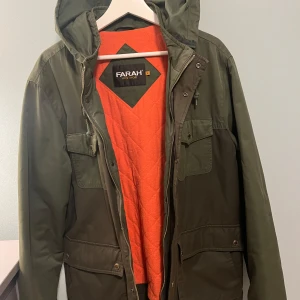 Grön parkajacka från Farah - Säljer en grön parkajacka från Farah Vintage med orange quiltat foder. Jackan har en huva och flera praktiska fickor. Perfekt för höst och vår med sin stilrena design och funktionella detaljer.
