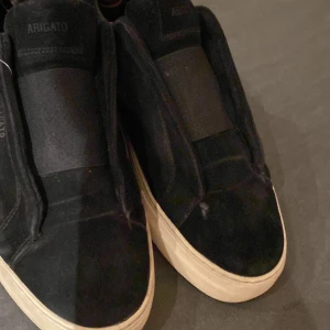 Svarta sneakers från Axel Arigato - Snygga svarta sneakers från Axel Arigato i mocka med elastiska paneler för enkel påtagning. Skorna har en vit sula som ger en stilren kontrast. Perfekta för en avslappnad och trendig look.