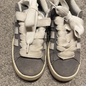 Grå Adidas Campus sneakers - Säljer ett par grå Adidas Campus sneakers med vita detaljer och snörning. Skorna har en klassisk design med de ikoniska tre ränderna på sidan. Perfekta för en avslappnad stil.Man får även gråa snören till 