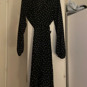 Svart prickig klänning - Snygg svart klänning med vita prickar och långa ärmar. Perfekt för en stilren look. Klänningen har en knytning i midjan som ger en fin passform.