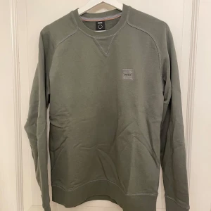 Boss Tröja - Säljer en stilren grön tröja från BOSS i storlek S. Tröjan har långa ärmar och en diskret logotyp på bröstet. Perfekt för en avslappnad look. Skick 10/10