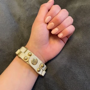 Beige armband med gulddetaljer - Snyggt beige armband med guldfärgade detaljer och nitar. Armbandet har en unik design med flätade band och tryckknappar för justering. Perfekt för att ge en stilren touch till din outfit.