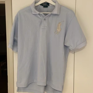 Ljusblå pikétröja från Ralph Lauren - Säljer en stilren ljusblå pikétröja från Ralph Lauren med klassisk krage och knappar. Tröjan har en broderad logotyp på bröstet och är kortärmad. Perfekt för en avslappnad men ändå elegant look.