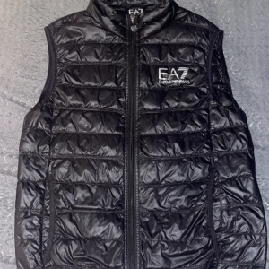 Armani Väst - Snygg svart dunväst från EA7 med dragkedja framtill. Västen har ett quiltat mönster och är perfekt för kyligare dagar. Den har en stilren design med logotyp på bröstet. Lite söndeig vid armen