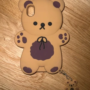 Brun björnformad mobilskal - Säljer ett gulligt björnformad mobilskal i brun färg med mörkare detaljer. Skalet har en liten björnberlock som hänger i en kedja. Perfekt för att ge din mobil en söt och unik look!🤍🌟