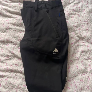 Svarta byxor från Burton - Snygga svarta byxor från Burton med dragkedja och logotypdetaljer. Perfekta för utomhusaktiviteter med en bekväm passform och praktiska fickor. Tillverkade i slitstarkt material.