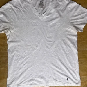 Vit v-ringad t-shirt från Ralph Lauren - Säljer en klassisk vit t-shirt från Ralph Lauren med v-ringning och korta ärmar. Den har en liten broderad logga i mörkblått på framsidan. Perfekt för en stilren och avslappnad look.